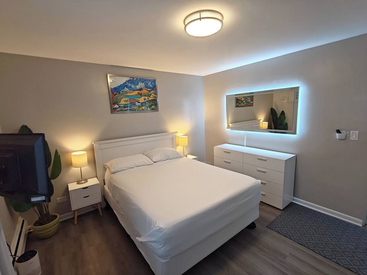 A bedroom with a bed, bedside tables, a dresser, a mirror, and a painting on the wall.