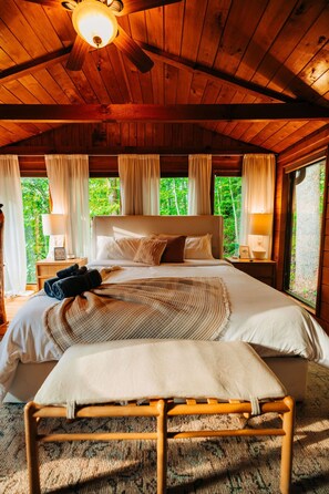 Ein Schlafzimmer einer Holzhütte mit Bett, Vorhängen, einem Nachttisch und Blick auf die grüne Landschaft.