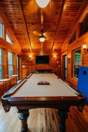 A pool table in a wooden room with a ceiling fan, windows, and a flat-screen TV mounted on the wall. - RRG cottage - cave fire pit, hot tub, & pool table (Stanton)