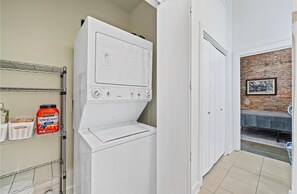 A laundry room with a white washer and dryer, a shelf with laundry detergent, and a bench in a brick-walled adjacent room.