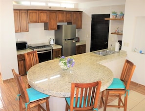 A kitchen with wooden cabinets, a granite countertop, a stainless steel refrigerator, and a dining area with wooden chairs and a table.