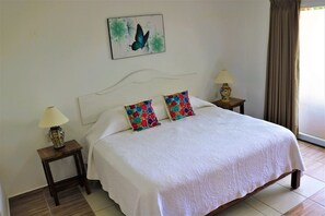 A neatly made bed with a white bedspread and two colorful pillows, a small bedside table with a lamp, and a wall-mounted artwork of a butterfly. - Sea-view condo w/ pool & palapa (Puerto Escondido)