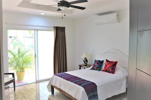 A hotel room with a bed, a ceiling fan, a window with curtains, a potted plant, and a view of palm trees.