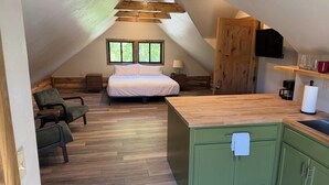 Een knus zolderkamer met een bed, een stoel, een houten plafond en een keukenhoek met een spoelbak en kasten.