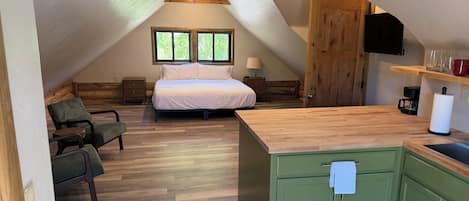 A cozy attic bedroom with a bed, a chair, a wooden ceiling, and a kitchen area with a sink and cabinets.