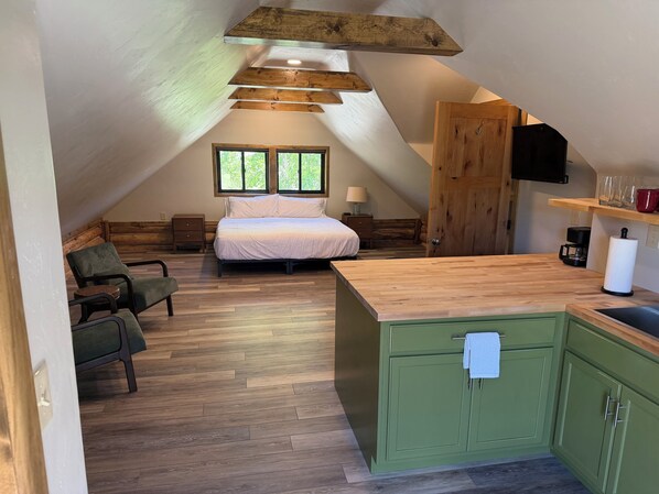 A cozy attic bedroom with a bed, a chair, a wooden ceiling, and a kitchen area with a sink and cabinets.