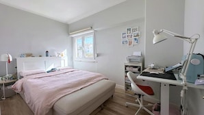 Quarto com cama, mesa de cabeceira, luminária, escrivaninha e janela.