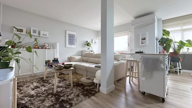 Uma sala de estar moderna com sofá, mesa de centro e tapete. Há uma área de cozinha com geladeira e micro-ondas.