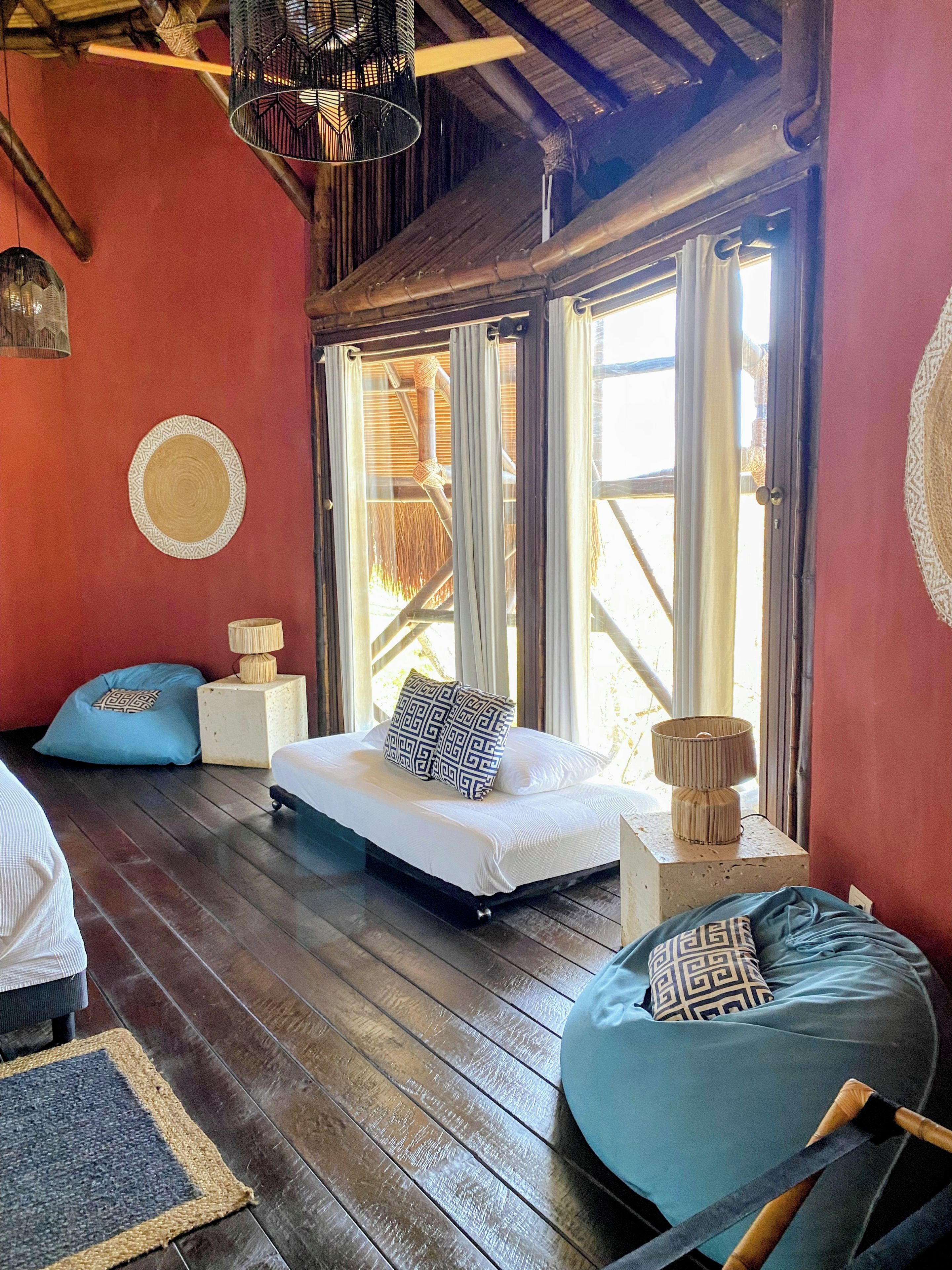 A room with a bed, a blue bean bag, a small table with a lamp, and a window with a view of bamboo.