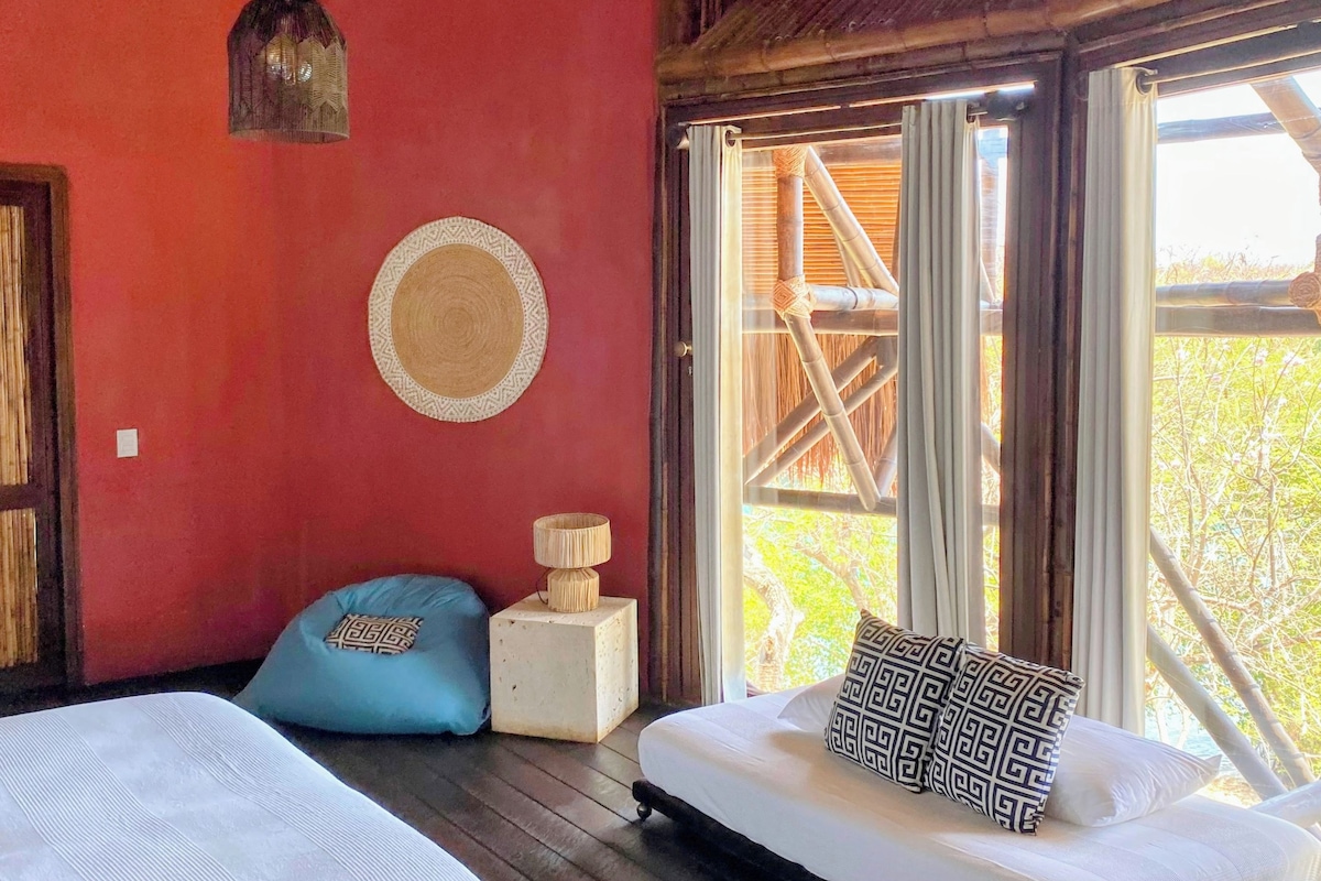 A room with two beds, a ceiling fan, a window with a view, and a woven lamp.