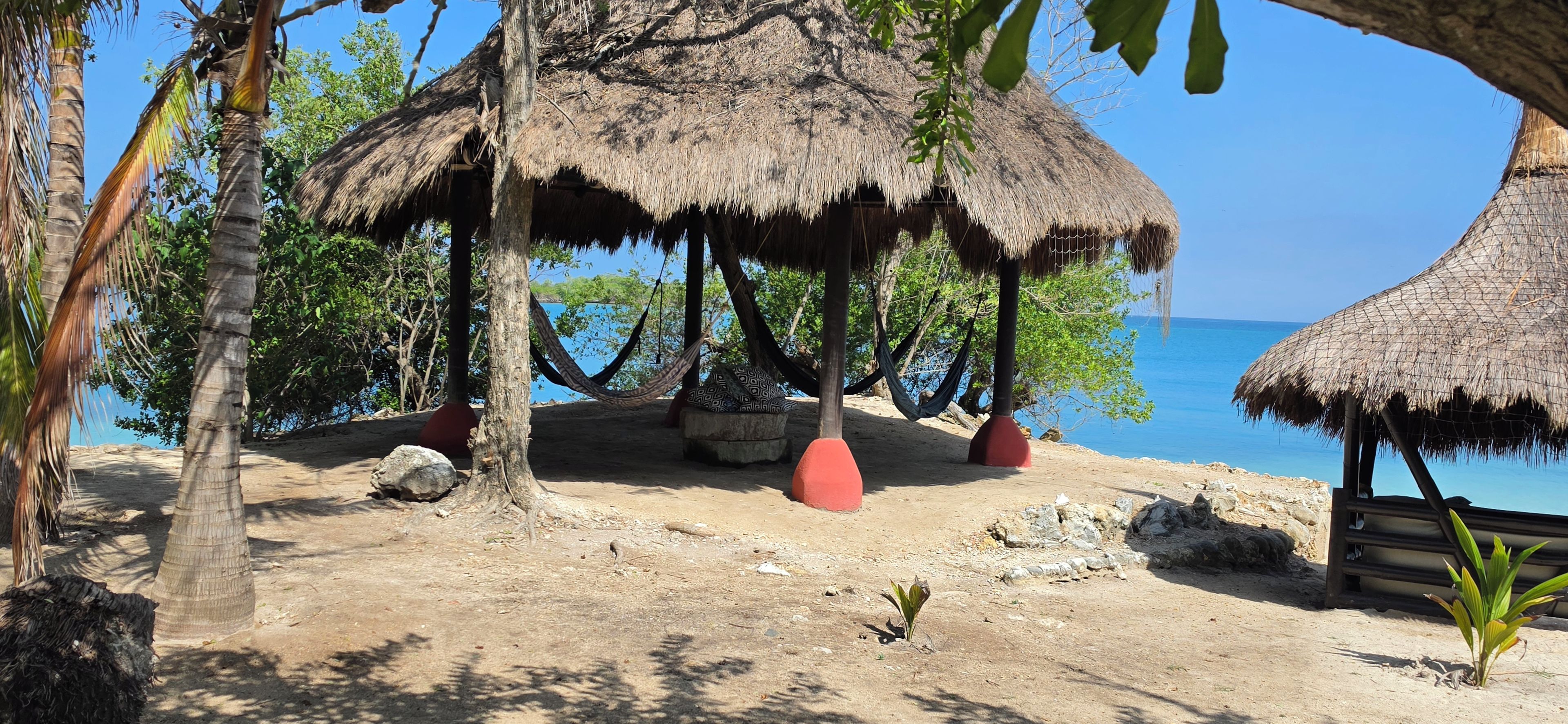 Escena de playa con cabañas de techo de paja, palmeras y cielo azul despejado.