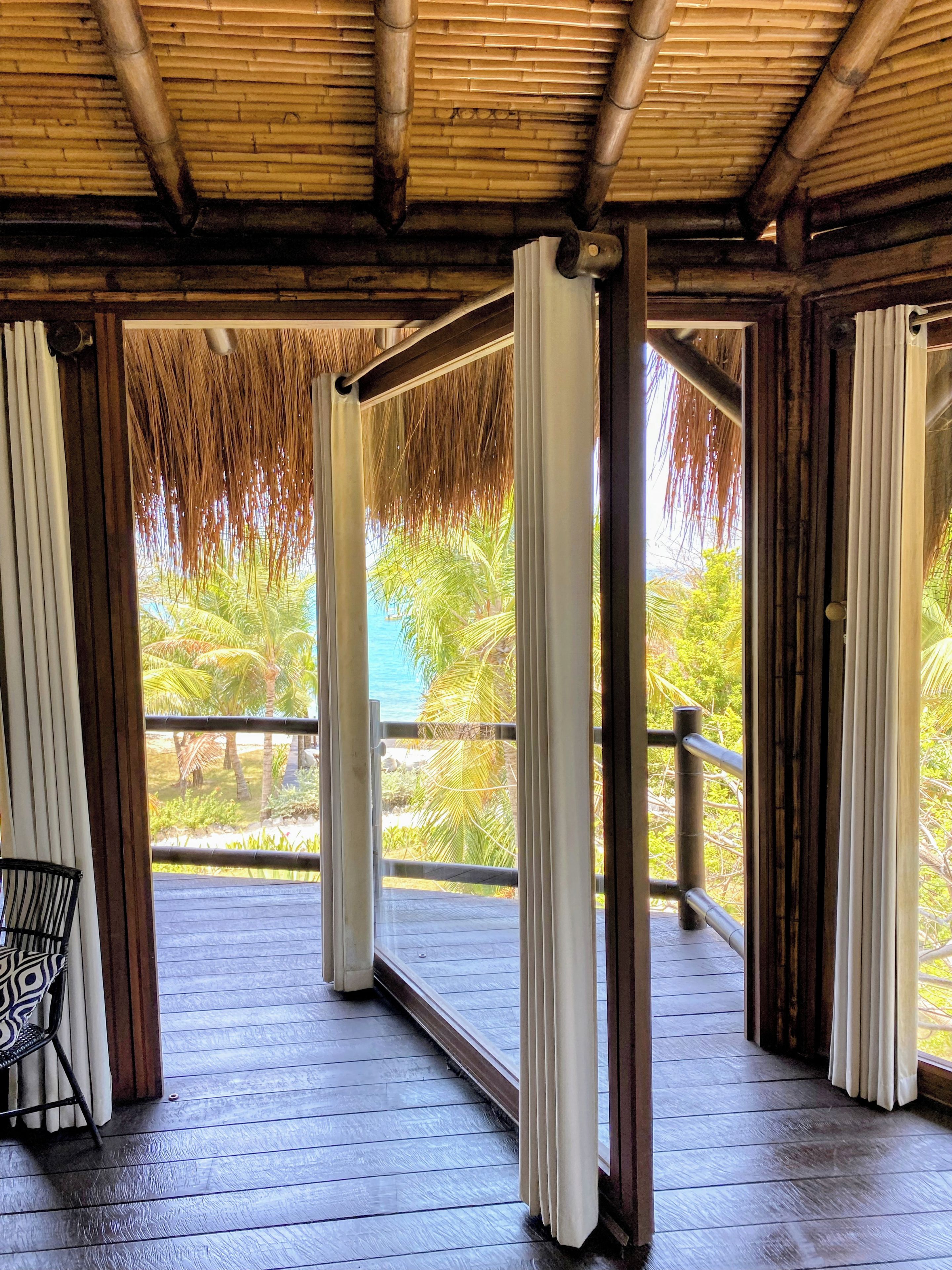 A covered outdoor area with a wooden floor, a chair, and a view of palm trees and water.