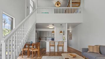 A spacious living area with a high ceiling, a dining table with chairs, a sofa, and a kitchen in the background.