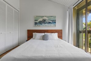 A bedroom with a large bed, a wooden headboard, a painting of a wave, and a view of greenery through a window.