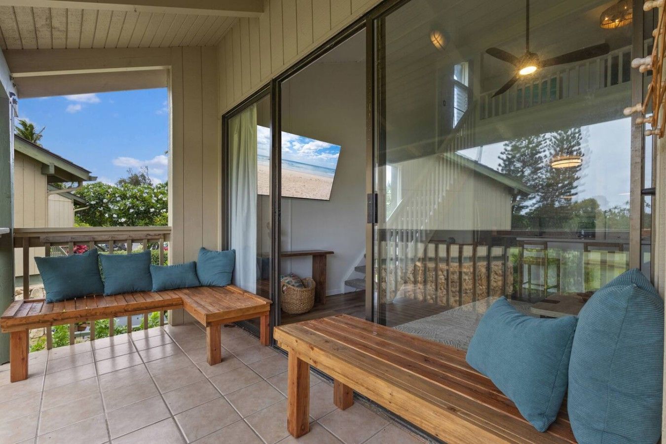 A covered outdoor seating area with wooden benches and cushions, a glass door leading to a balcony, and a view of the beach.
