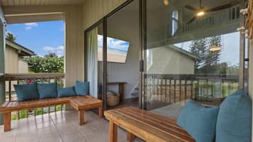 A covered outdoor seating area with wooden benches and cushions, a glass door leading to a balcony, and a view of the beach.
