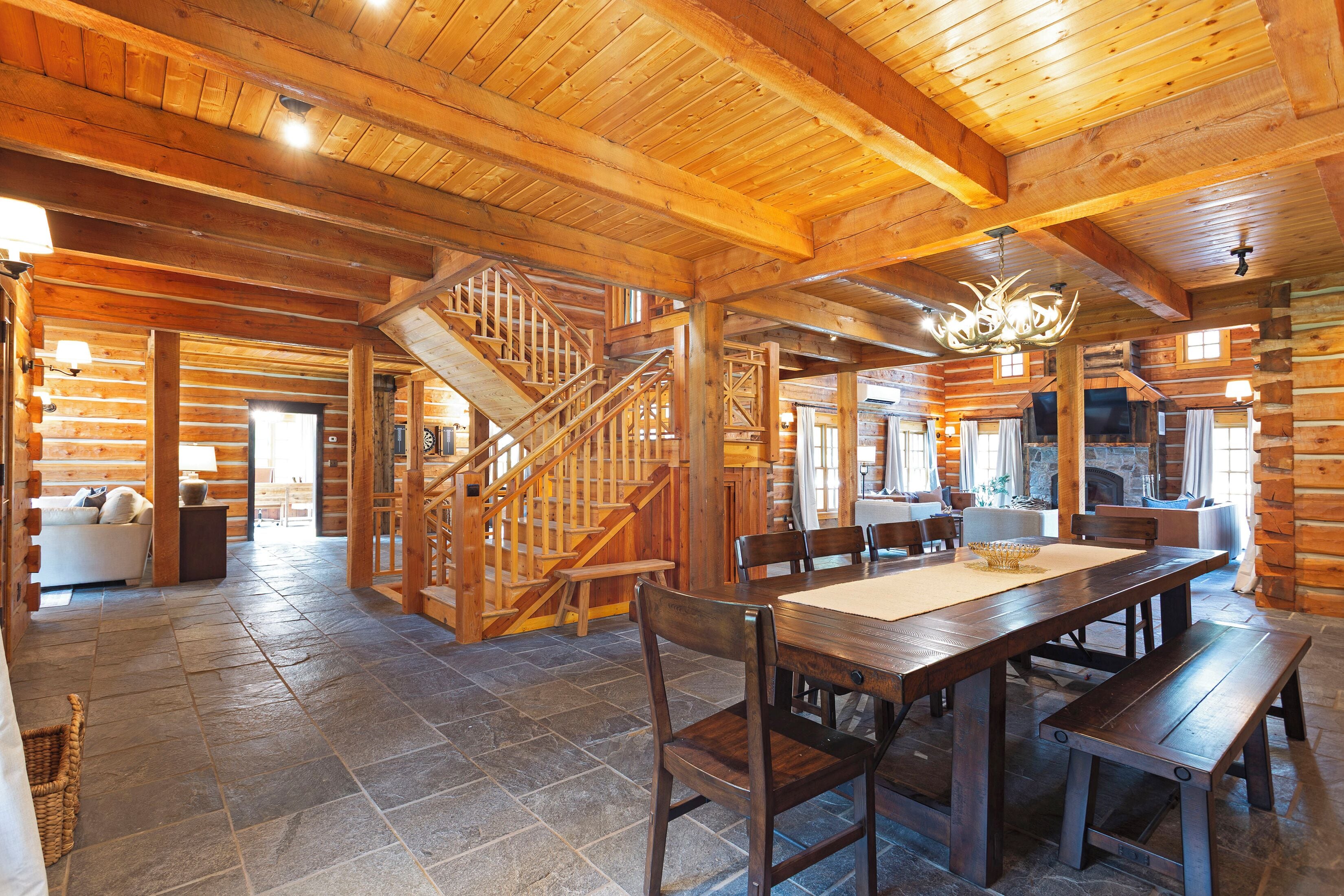 A log cabin interior with a stone floor, wooden staircase, dining area, and a chandelier.