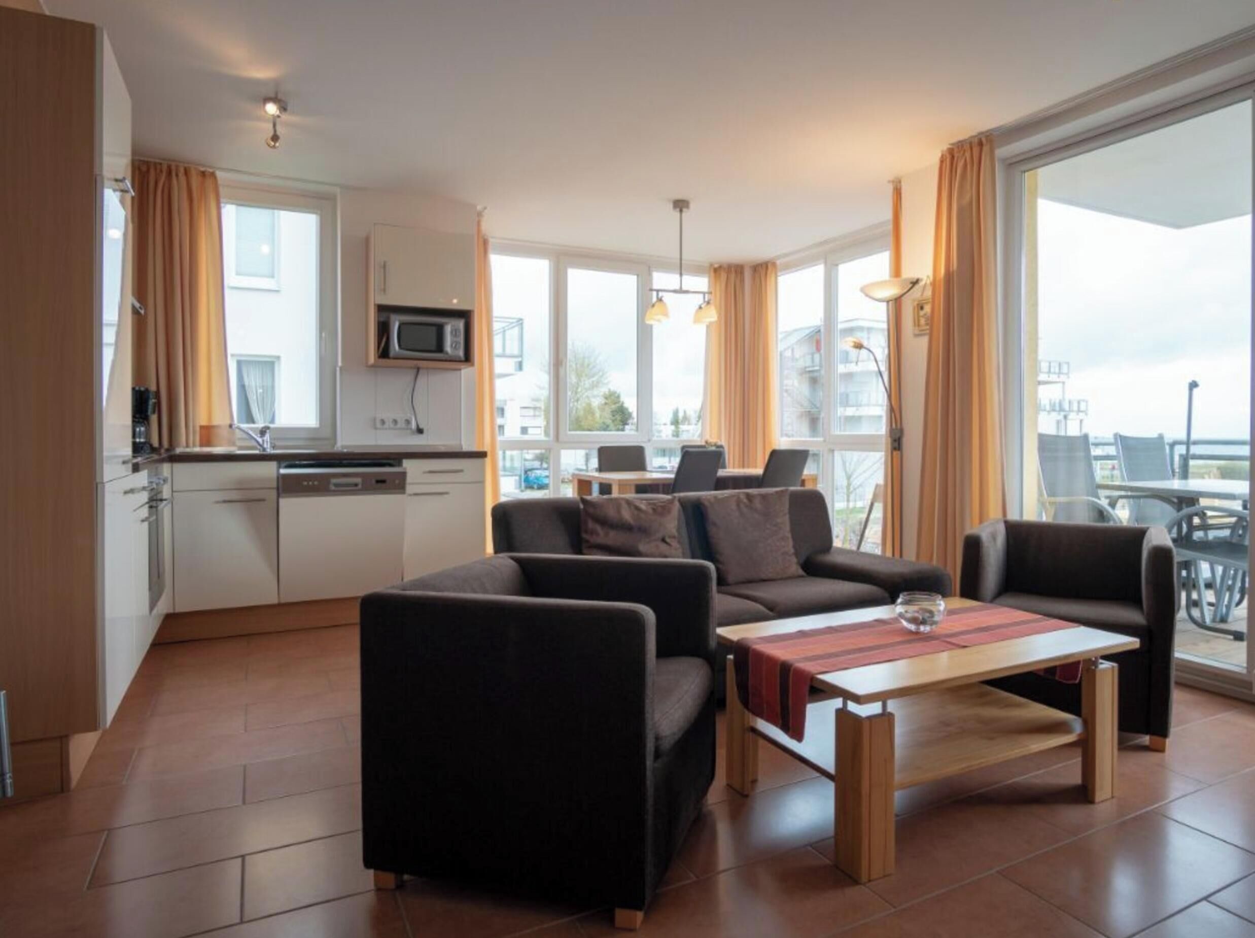 A modern living room with a sofa, armchairs, and a coffee table. There is a kitchen area with a microwave and cabinets. Large windows provide natural light and a view of the outside.