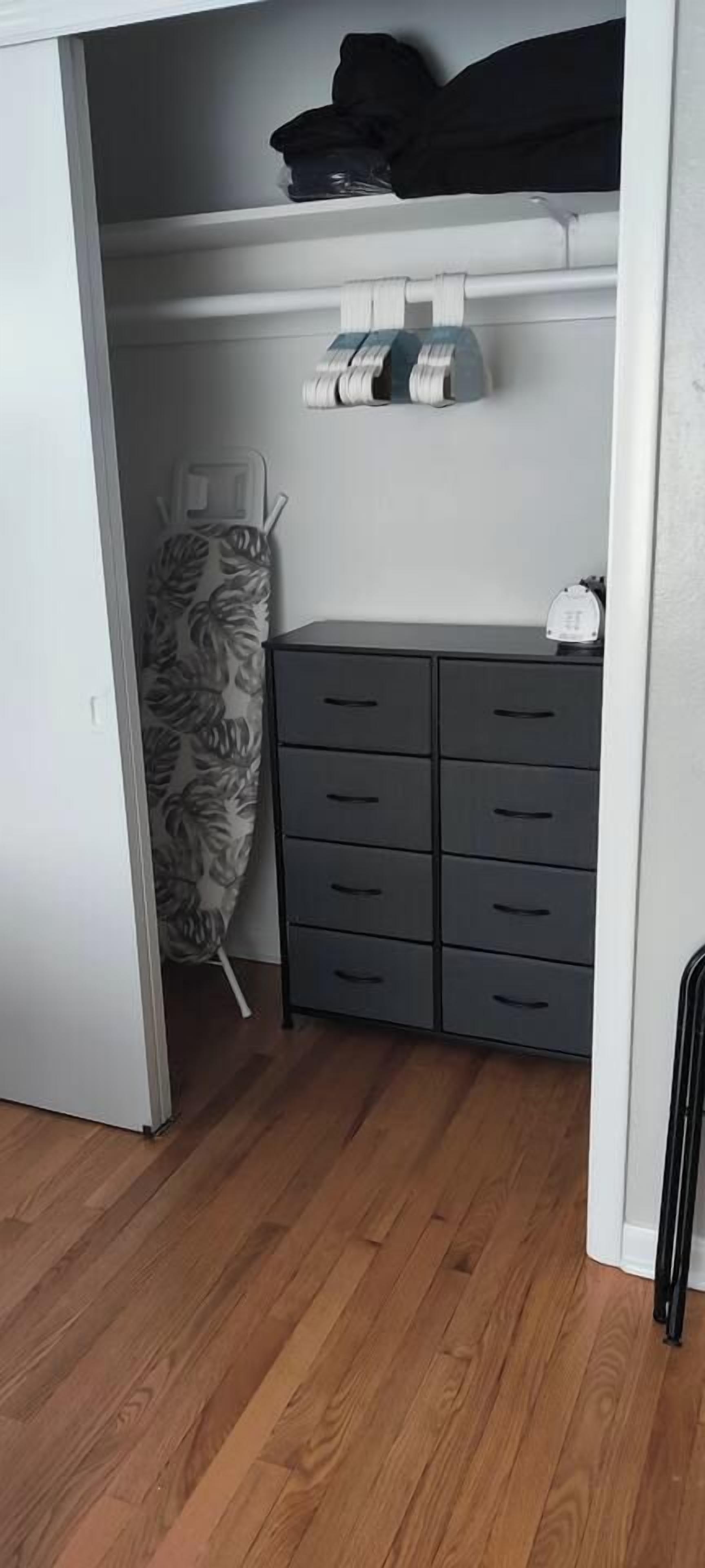 A room with a wooden floor, a grey storage unit with multiple drawers, and a patterned ironing board.
