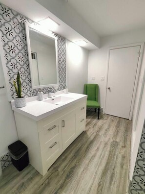 A bathroom with a white vanity, a large mirror, a green chair, and patterned wallpaper. - The Rooftop - Haven! (Waukegan)