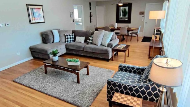 A living room with a sectional sofa, a patterned armchair, a coffee table, and a lamp. - The Rooftop - Haven! (Waukegan)