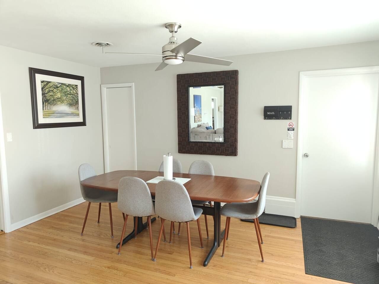 A dining area with a wooden table, grey chairs, a ceiling fan, a framed picture, a mirror, and a mail slot.