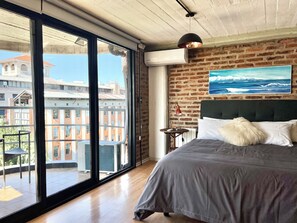 Ett sovrum med ett stort fönster, en säng, en sänggavel, en väggmonterad luftkonditioneringsanläggning och en målning som föreställer ett havsskepp.