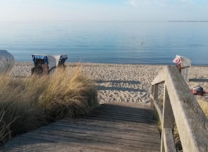 Ein hölzerner Steg führt zu einem Sandstrand, im Hintergrund liegt ruhiges Wasser.