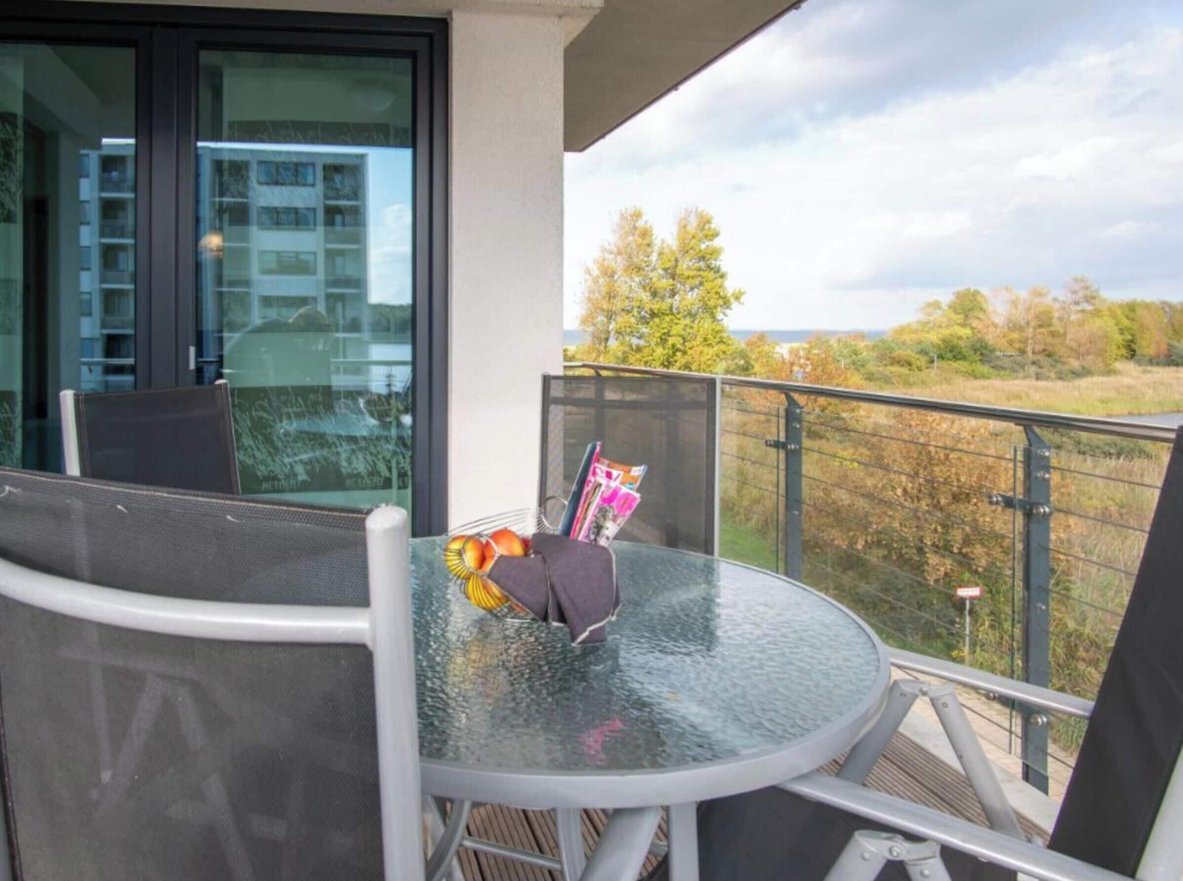 Ein Tisch mit Glasplatte, ein Stuhl und eine Fruchtkorb auf einem Balkon mit Blick auf Bäume und Gebäude.
