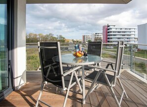 Ein Balkon mit einem Glastisch und Stühlen, einer Obstschale und Blick auf Gebäude und Bäume.