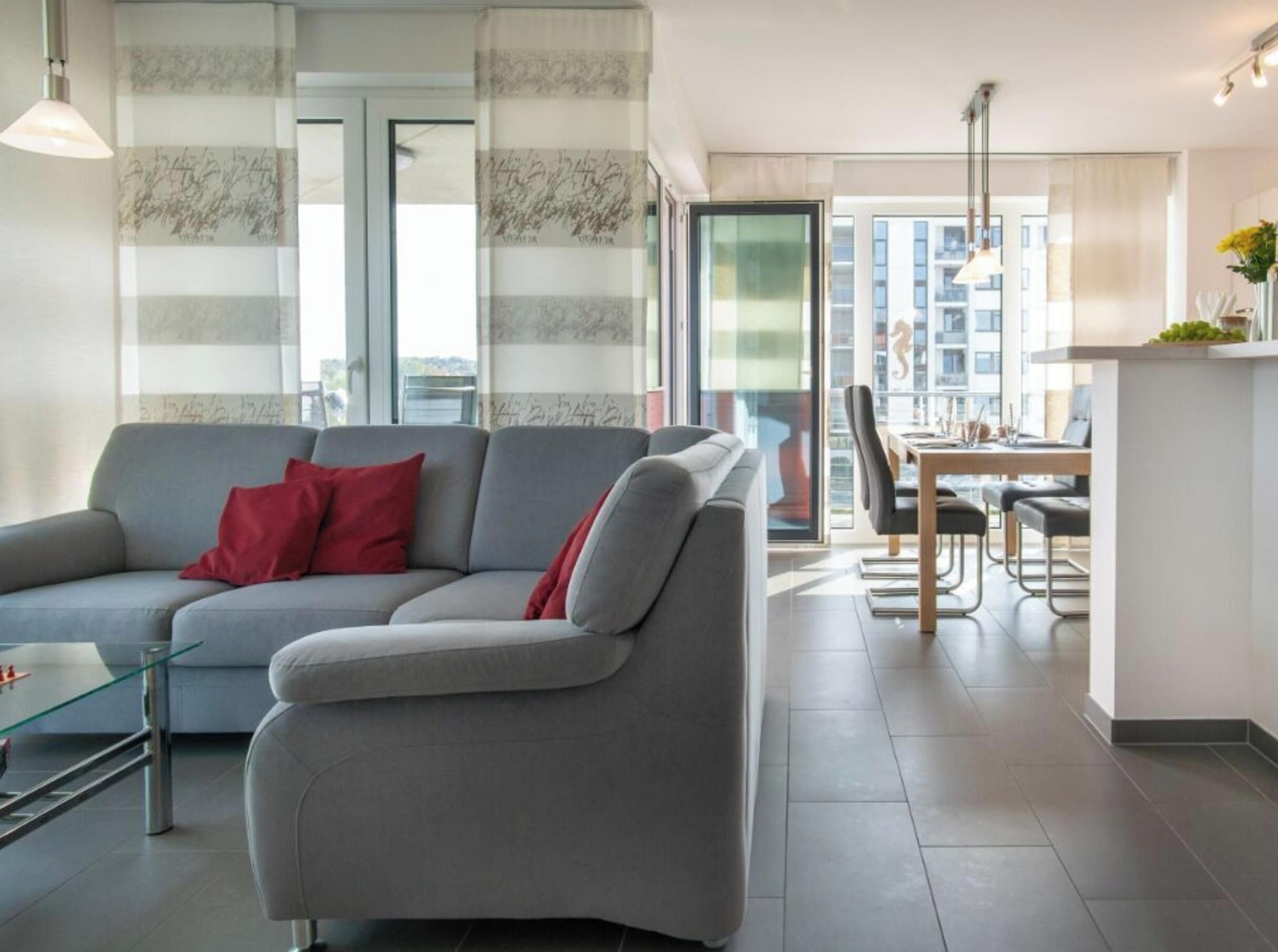Uma sala de estar moderna com um sofá cinzento, almofadas vermelhas, uma mesa de centro de vidro e um espaço de jantar com uma mesa e cadeiras.