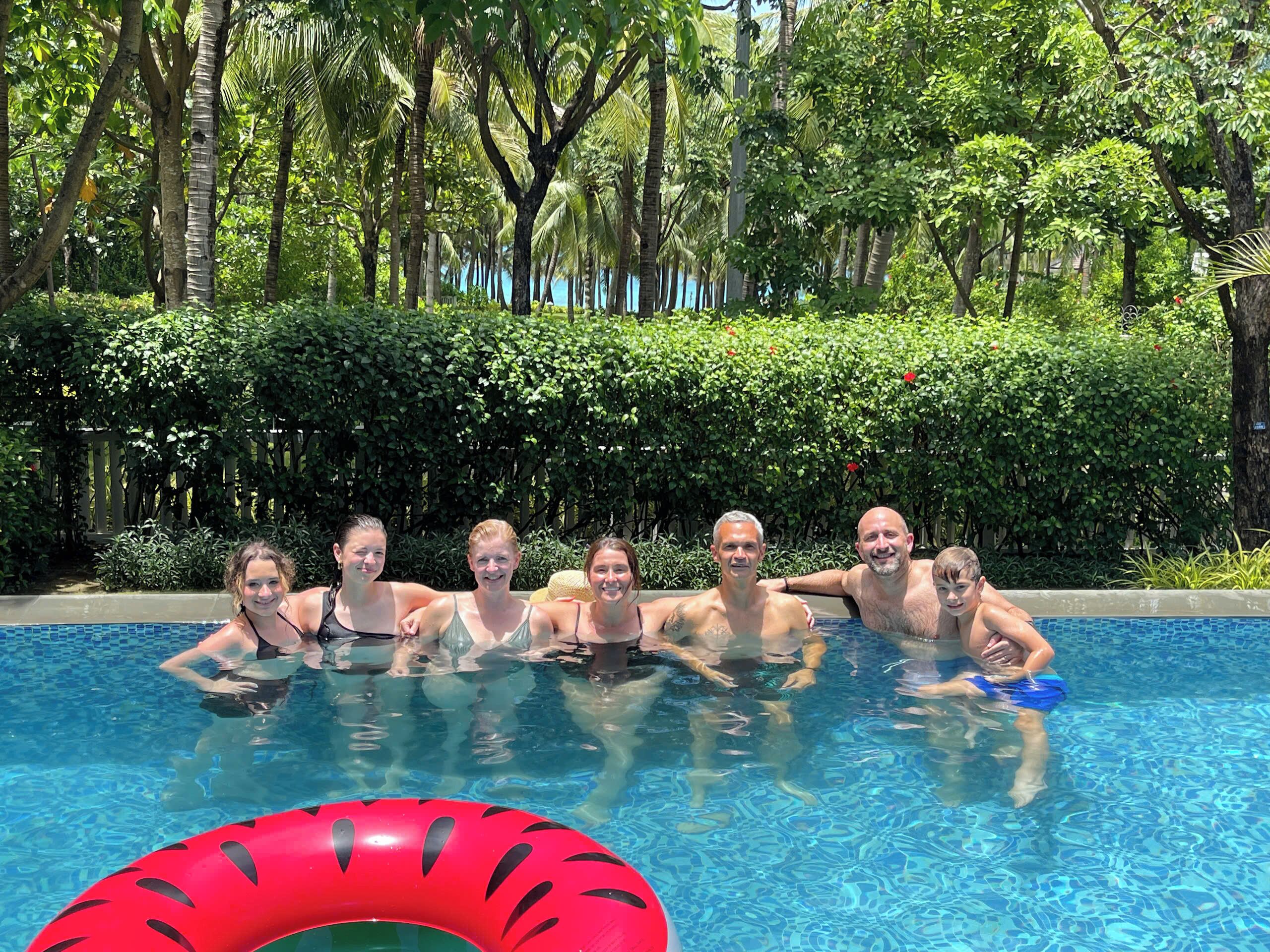 A group of people in a swimming pool with a red inflatable ring, surrounded by lush greenery.