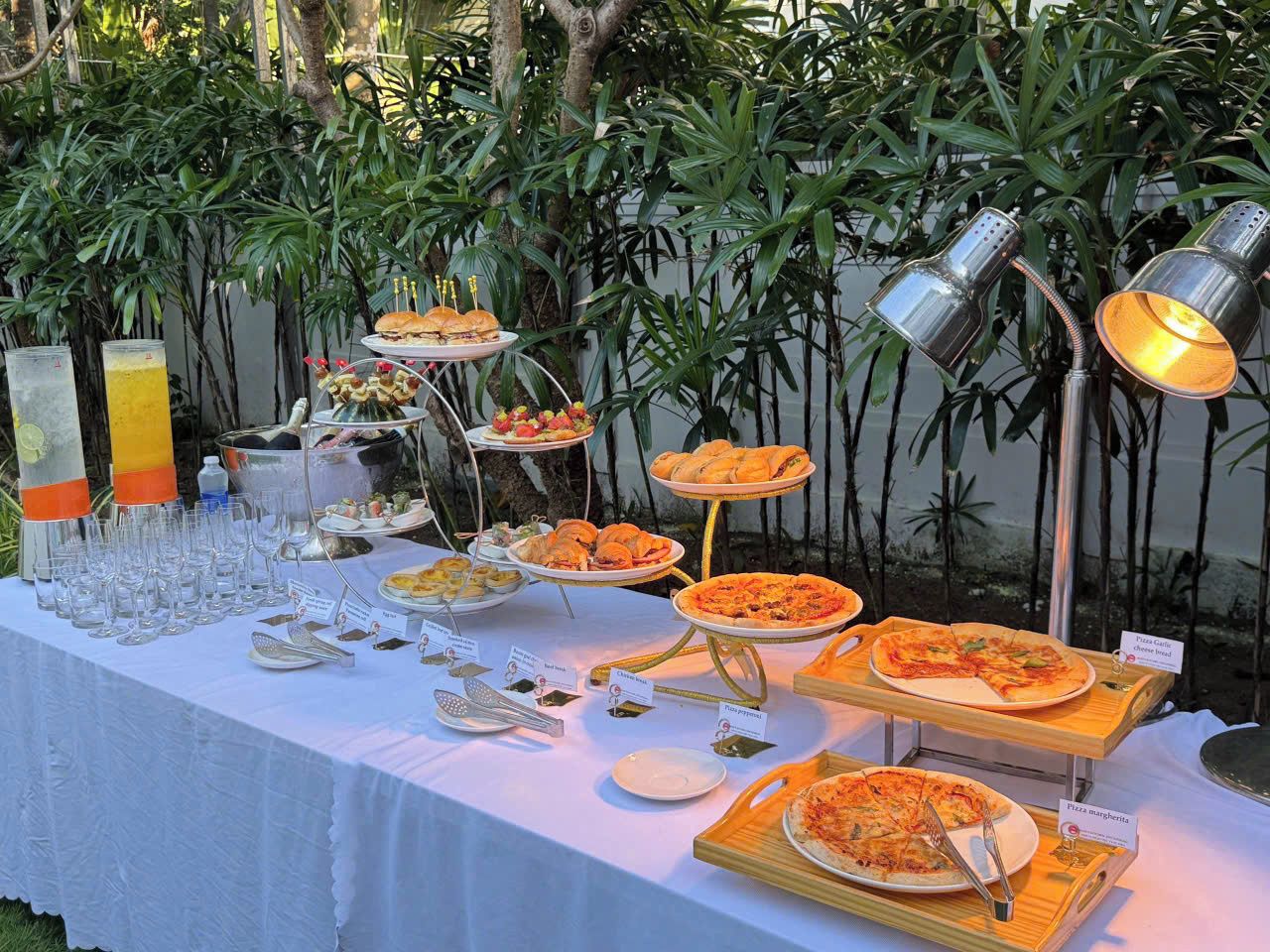 A buffet table with various food items and utensils, set against a backdrop of greenery.