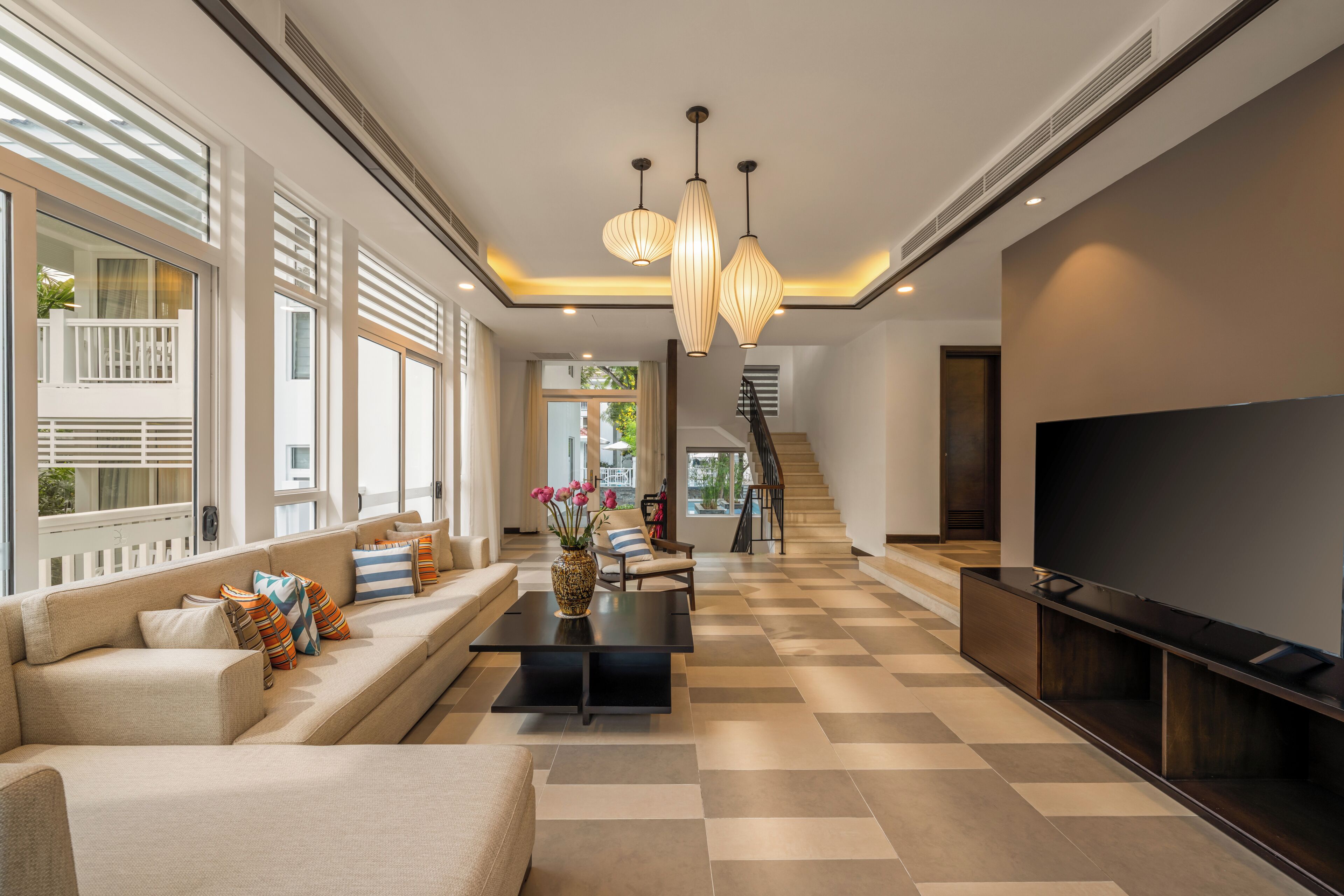 A modern living room with a sofa, coffee table, and TV stand, a staircase, and large windows.