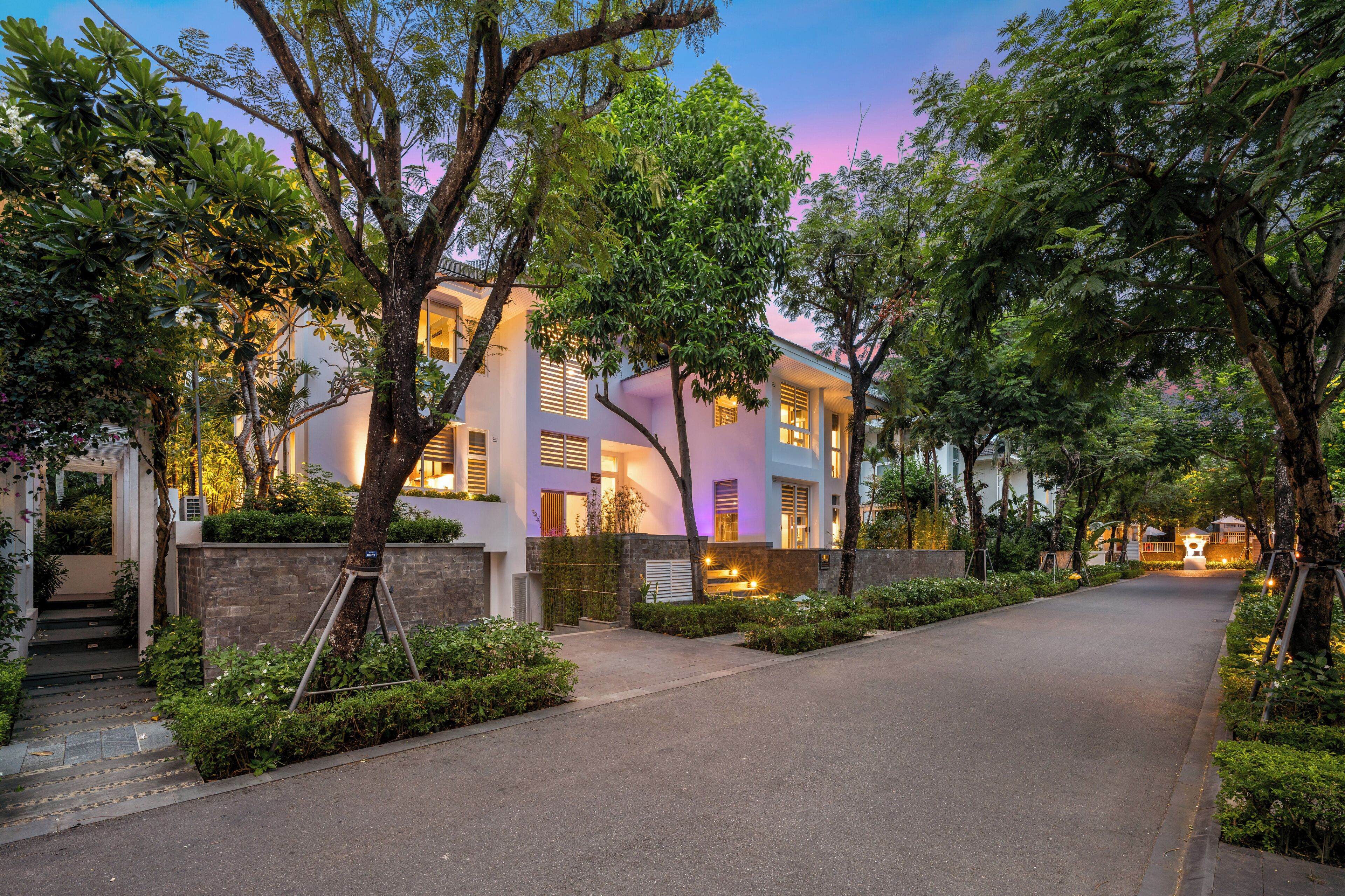 A modern two-story building with a paved driveway, surrounded by lush greenery and trees.