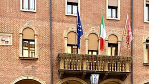 Un bâtiment en briques rouges, doté de nombreuses fenêtres et d’un balcon orné de drapeaux.