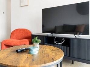 Un salon moderne avec une table ronde en bois, une télécommande, une petite plante en pot et un meuble noir pour appareils électroniques, sur lequel est posée une télévision à écran plat.