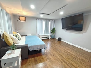 Quarto moderno com cama, mesa de cabeceira, abajur e televisão na parede.