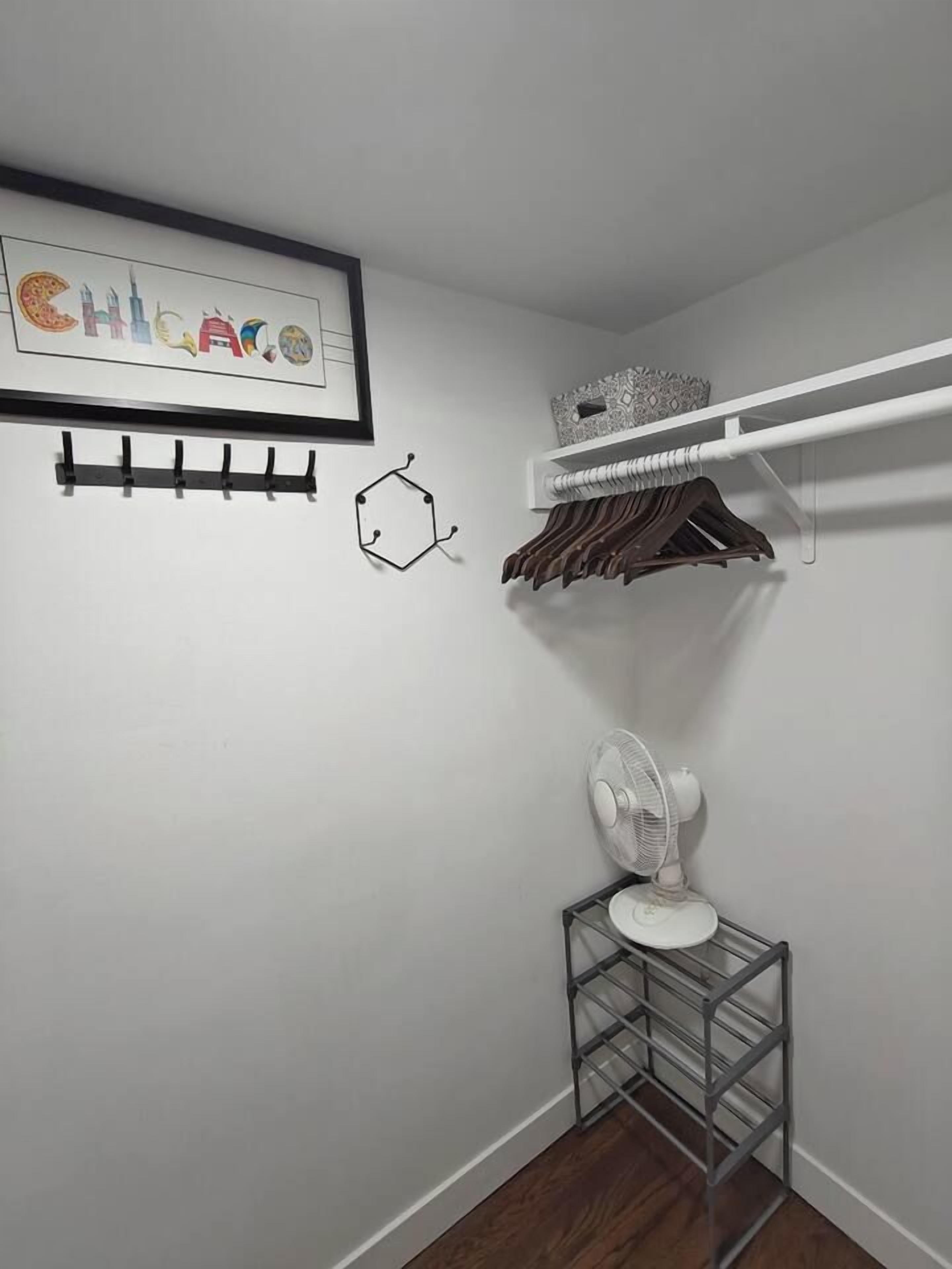 Un rincón de una habitación con una pared blanca, un cuadro enmarcado, un ventilador blanco y un perchero de metal.