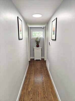 A hallway with a white door at the end, a potted plant on a white console table, and framed pictures on the walls.
