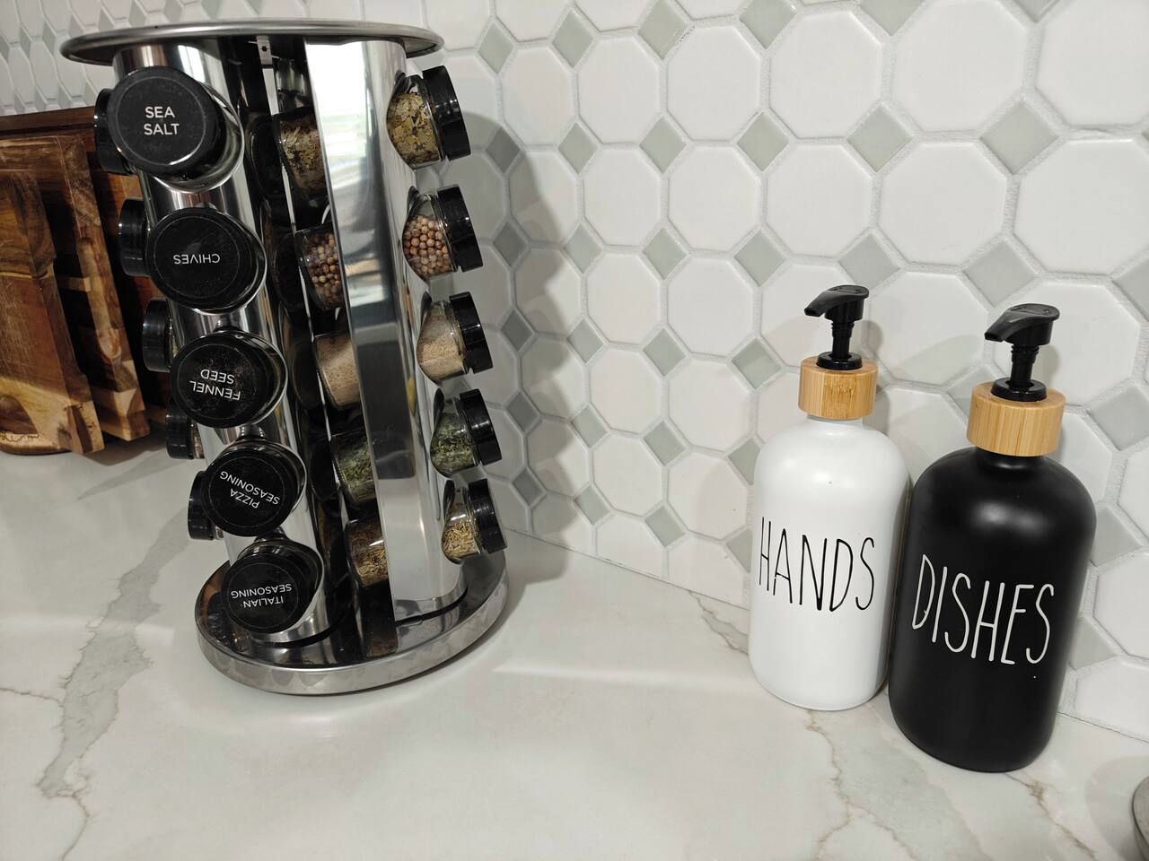 A kitchen counter with a spice rack, two labeled bottles, and a hexagonal tiled backsplash.