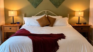Ein ordentlich bezogenes Bett mit weißer Daunendecke und zwei Kissen, an beiden Seiten mit Nachttischen mit Lampen, in einem Zimmer mit Deckenventilator und zwei Wanddekorationen.