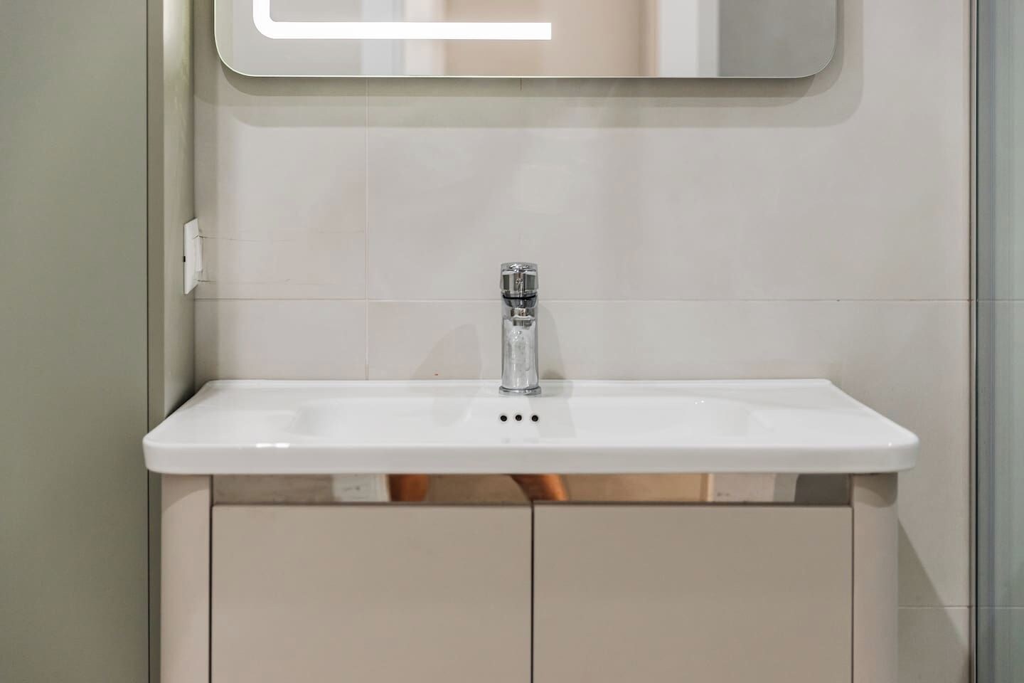 A bathroom with a white sink, a mirror above it, and a plain tiled wall.