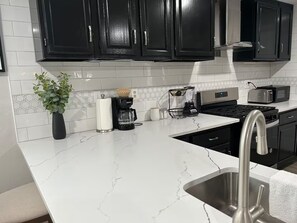 A modern kitchen with black cabinets, a white marble countertop, a stainless steel sink, a coffee maker, a toaster, and a microwave. - Charming Country Home! (Azle)