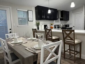 A dining area with a wooden table set for four, white chairs, and a kitchen with black cabinets and a white countertop. - Charming Country Home! (Azle)
