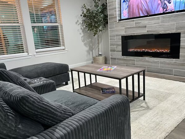 A modern living room with a sectional sofa, a coffee table, and a flat-screen TV mounted on a wall with a fireplace below. - Charming Country Home! (Azle)