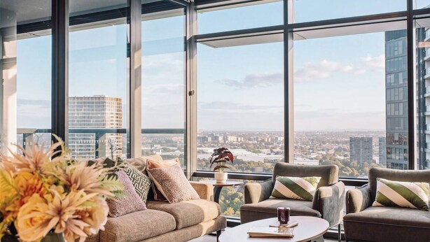 Uma sala de estar moderna com sofá, poltronas e uma mesa de centro, com vista para a cidade através de amplas janelas.