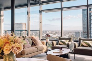 A modern lounge with a sofa, armchairs, and a coffee table, featuring a city view through large windows. - The Sky Perch 2- Walking distance to Accor stadium and Qudos bank arena (Sydney Olympic Park)