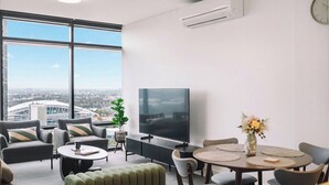 Uma sala de estar moderna com uma televisão grande, assentos confortáveis, uma área de refeições e vista para o horizonte urbano através de grandes janelas.