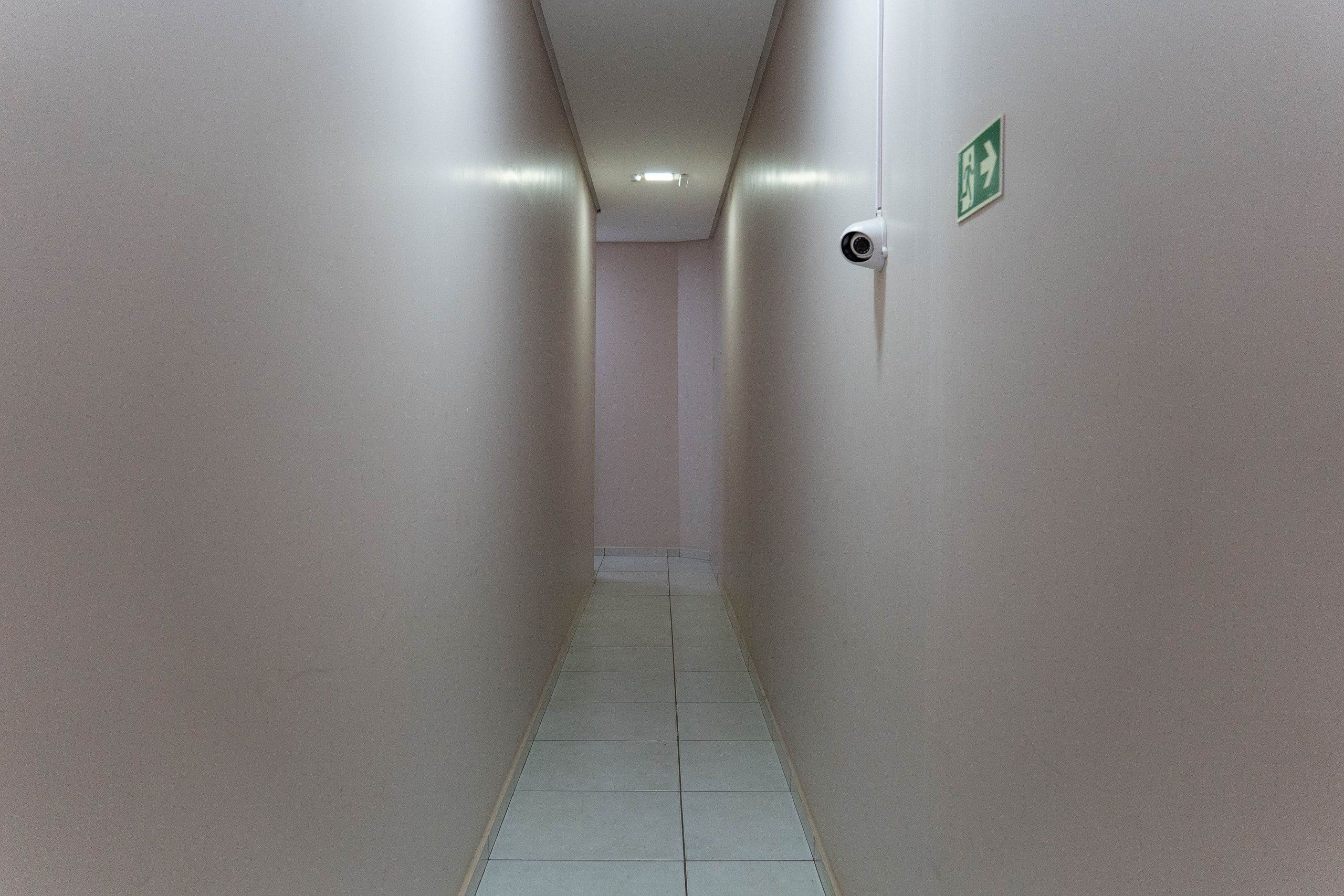 Un couloir étroit et bien éclairé, doté d’un sol carrelé, d’une caméra de surveillance et d’un panneau indiquant la sortie de secours sur le mur.